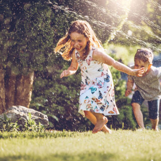 Wasserspiele für Kinder – Ein Mädchen und ein junge rennen durch die Wasserstrahlen eines Rasensprinklers.