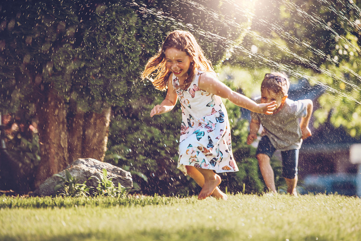 Wasserspiele für Kinder – Ein Mädchen und ein junge rennen durch die Wasserstrahlen eines Rasensprinklers.