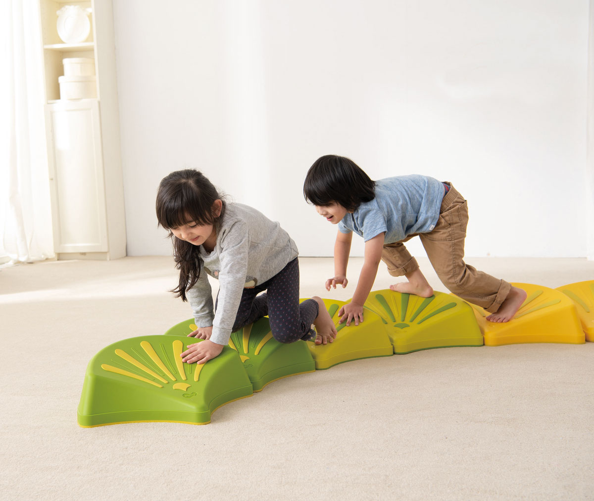 Bewegungsräume – Ein Mädchen und ein Junge klettern über einen Weg aus bunten Plastikmuscheln am Boden.