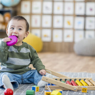 Objektspiel – ein Kleiner Junge sitzt mit seinem Spielzeug am Boden und beißt auf einen pinken Holzring.