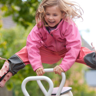 Bewegungspädagogik – Lachendes Mädchen in pinker Regenjacke und Gummistiefeln auf einer Wippe.