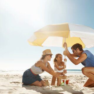 Sandkasten mit Sonnensegel – Mutter, Vater und Tochter bauen am Strand einen Sonnenschirm auf.