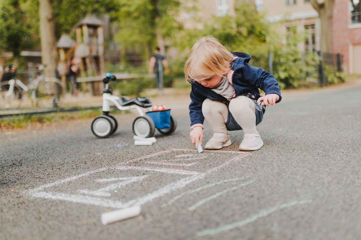 Kinderfreundliche Stadt – Ein kleines, blondes Mädchen malt mit Kreide auf die Straße.