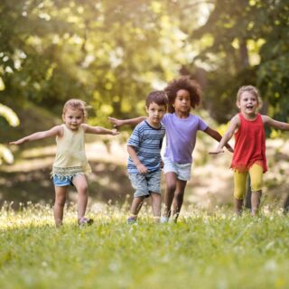Gruppenspiele draußen – Fünf Kinder, die in Sommerkleidung über eine Wiese rennen.