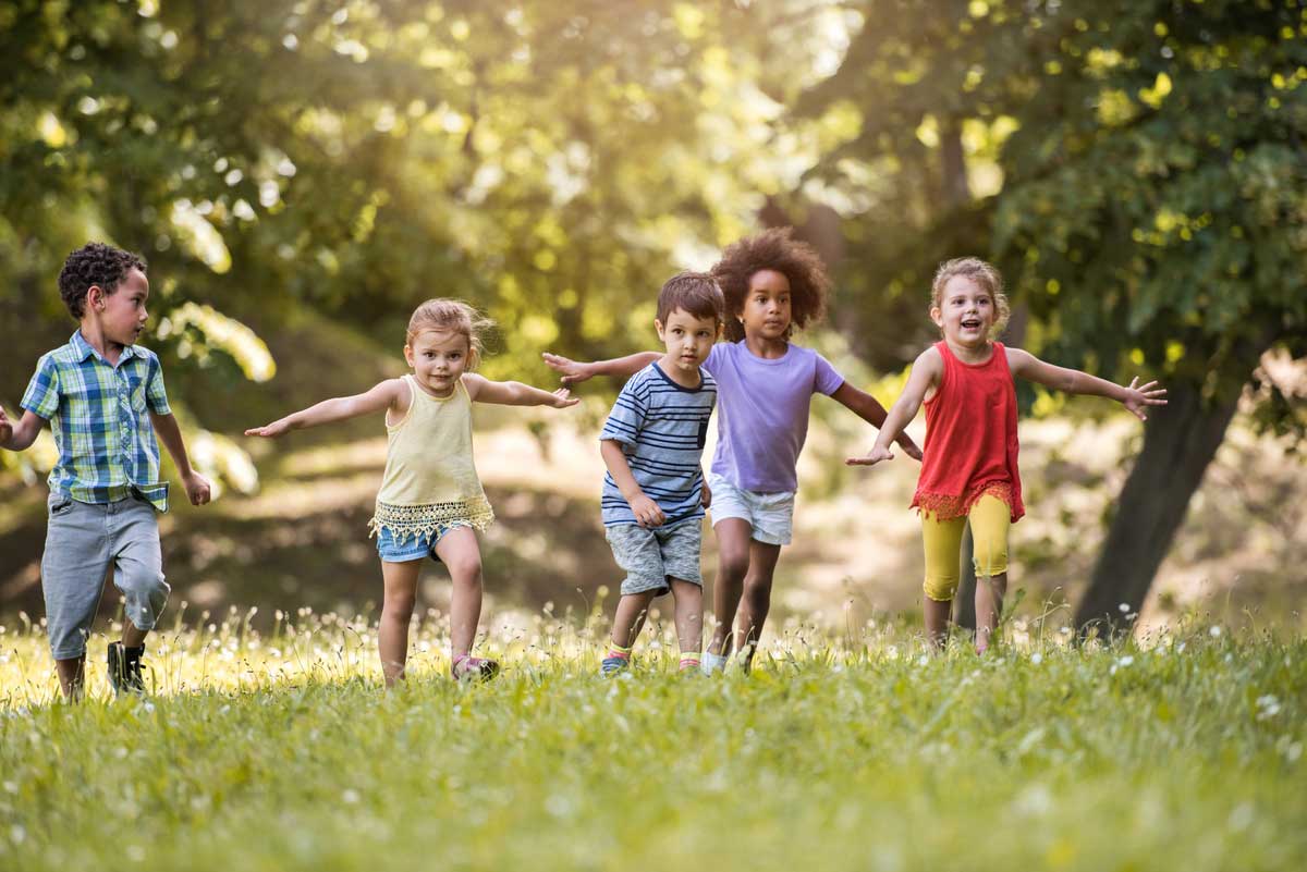 Gruppenspiele draußen – Fünf Kinder, die in Sommerkleidung über eine Wiese rennen.
