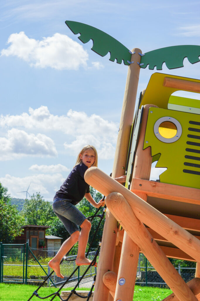 Ein blonder Junge klettert von der Spielanlage Geländewagen auf einem Themenspielplatz von eibe.