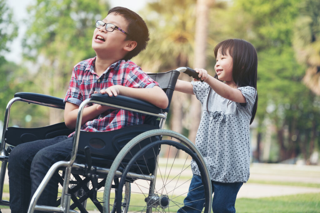 DIN 18040 barrierefreies Bauen von Spielplätzen – Ein lachendes Mädchen schiebt einen lachenden Jungen im Rollstuhl.