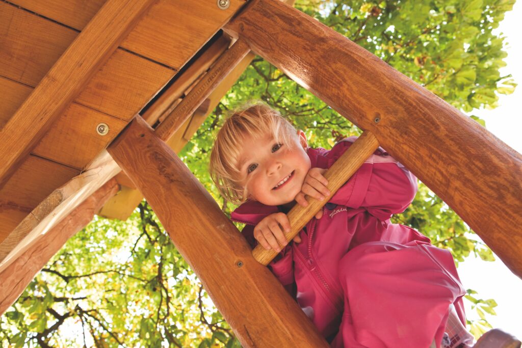 Farbwahrnehmung bei Kindern – Ein kleines Mädchen klettert die hölzerne Leiter einer Spielanlage hinauf und schaut dabei durch die Sprossen.