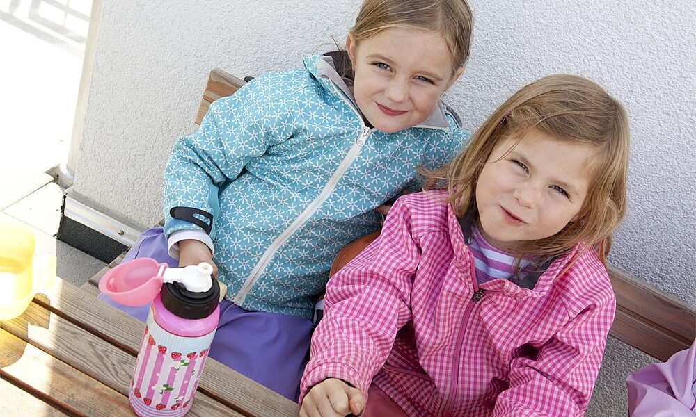Ruhepausen für Kinder – Zwei Mädchen in gemusterten Jacken sitzen auf einer Holzbank, an einem Holztisch, auf dem eine Trinkflasche steht.