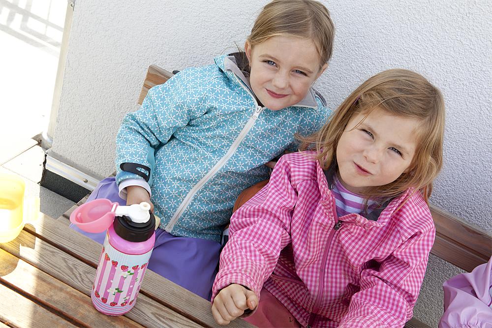 Ruhepausen für Kinder – Zwei Mädchen in gemusterten Jacken sitzen auf einer Holzbank, an einem Holztisch, auf dem eine Trinkflasche steht.