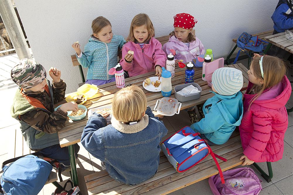 Ruhepausen für Kinder – Sieben Kinder in bunten Übergangsjacken machen Brotzeit in einer Kindersitzgruppe aus Holz.