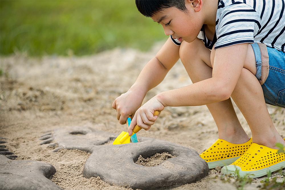 Spielplatzgestaltung – Ein Junge mit gelben Schuhen hockt im Sandkasten und buddelt eine Bodenplatte in Form eines Fossils aus.