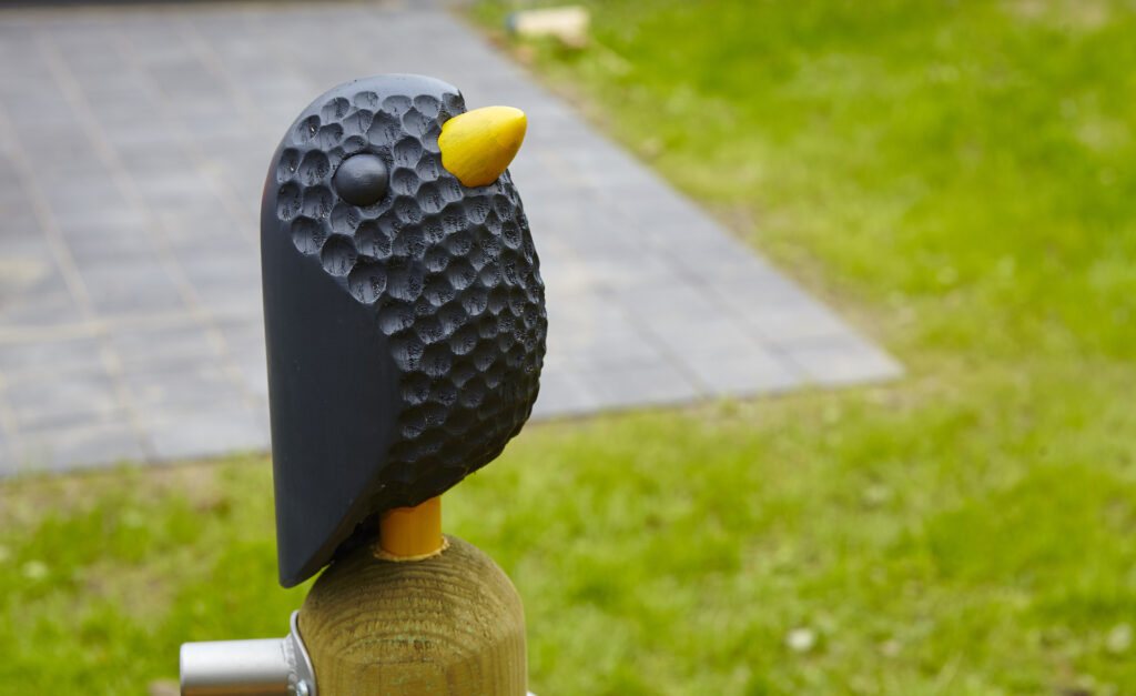Spielplatzgestaltung – Ein kleiner schwarzer Vogel aus Holz, der auf dem Pfosten einer Spielanlage sitzt.