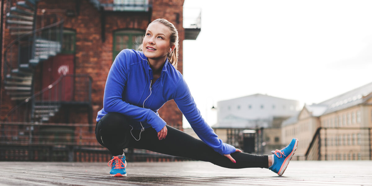 Sport in der Stadt – Eine junge Frau in blauer Trainingsjacke deht ihr Bein vor srädtischer Kulisse.
