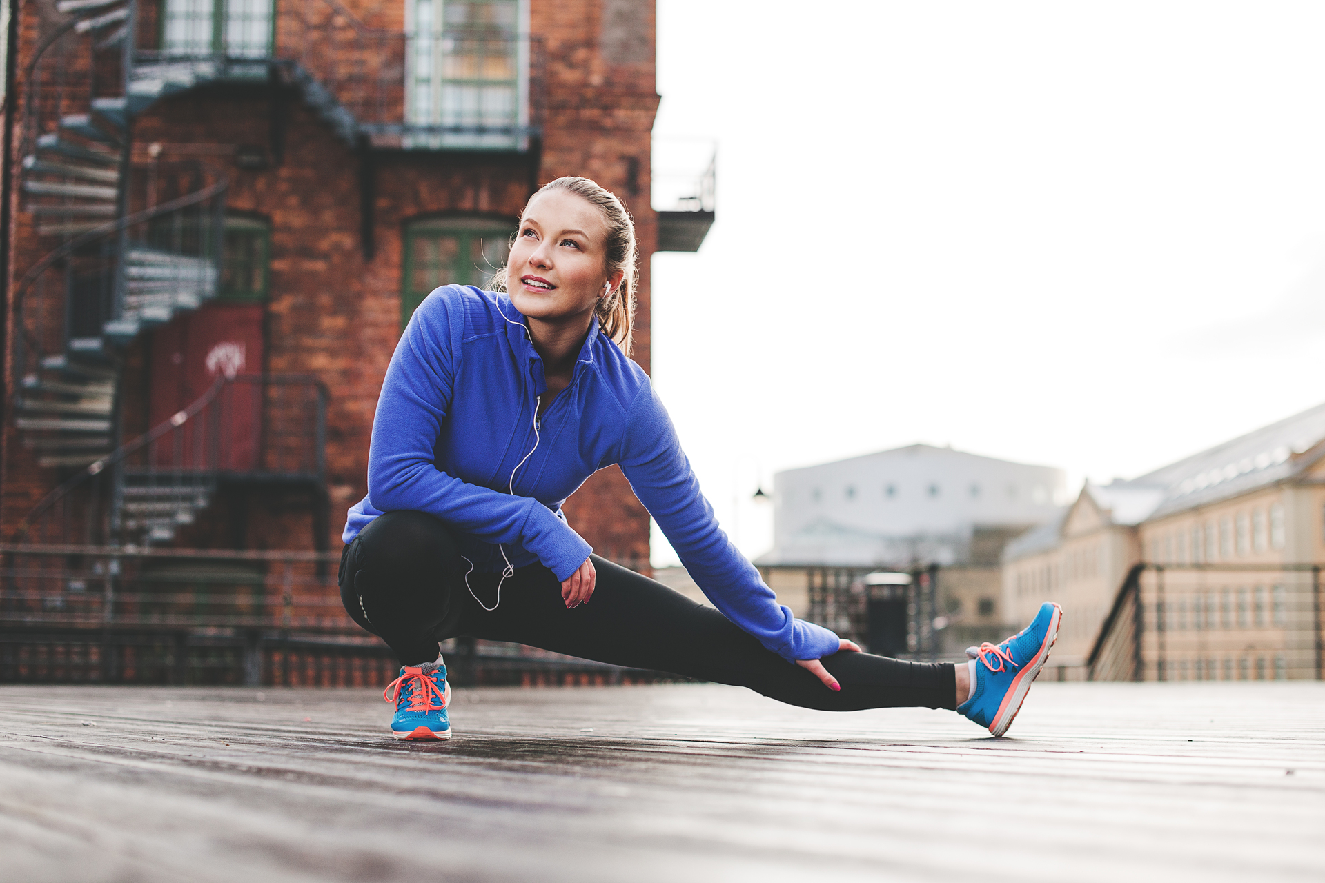 Sport in der Stadt – Eine junge Frau in blauer Trainingsjacke deht ihr Bein vor srädtischer Kulisse.