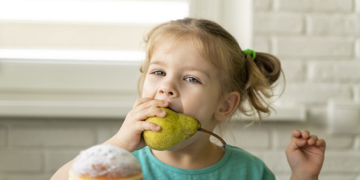 Übergewicht bei Kindern – Ein kleines, blondes Mädchen beißt in eine Birne, während vor ihr auf dem Tisch ein Krapfen mit Puderzucker liegt.
