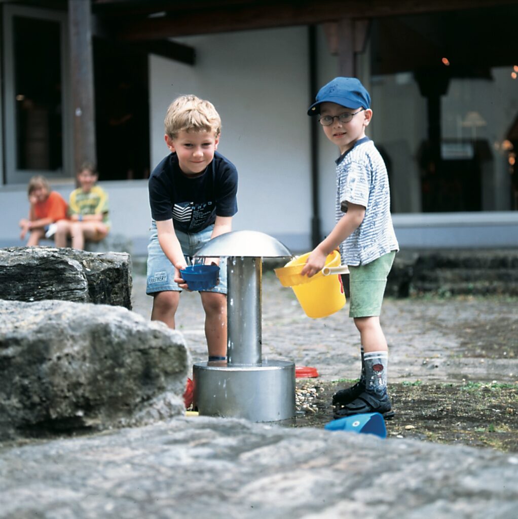 Zwei kleine Jungen stehen mit Eimern vor einem pilzförmigen Brunnen auf einem Wasserspielplatz.