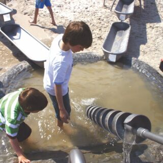 Zwei Jungen spielen auf einem Wasserspielplatz. Sie stehen in einer mit Wasser gefüllten Grube und blicken auf ein spiralförmiges Spielgerät; Ansicht von schräg oben.